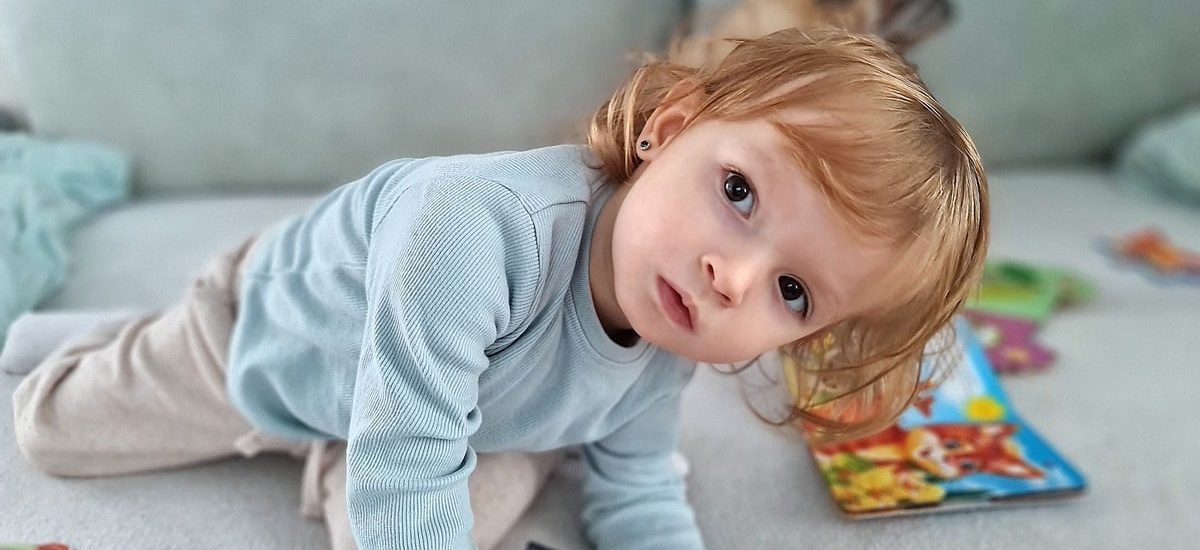 Adorable child kneels on a sofa, coloring with a pencil surrounded by toys in a cozy indoor setting.