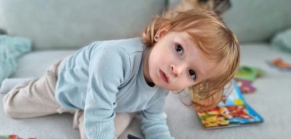 Adorable child kneels on a sofa, coloring with a pencil surrounded by toys in a cozy indoor setting.