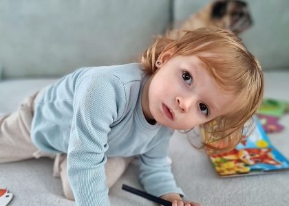 Adorable child kneels on a sofa, coloring with a pencil surrounded by toys in a cozy indoor setting.